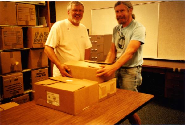 Image of two men working in a storage room with packing boxes on shelving visible behind them 